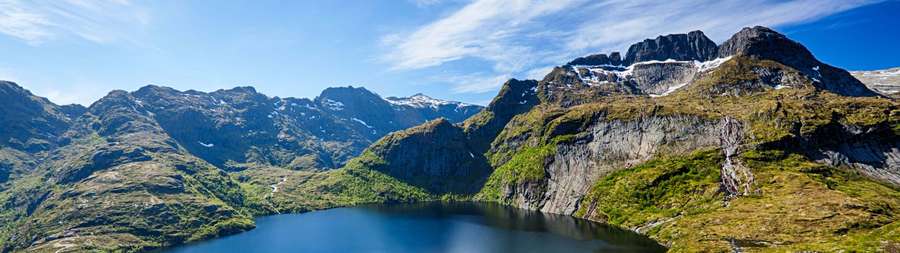 A calm mountain lake surrounded by green and snow-capped peaks under a clear blue sky. — 3840X1080 Background