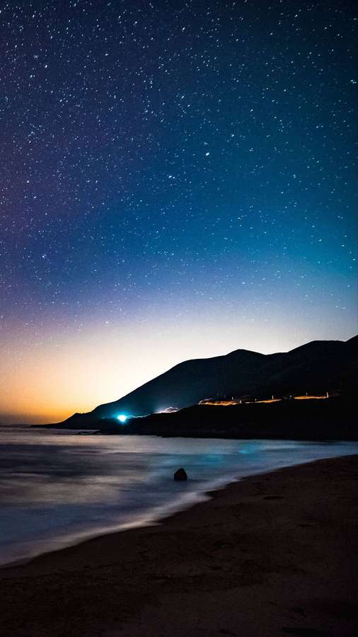 Starry night over a beach with hills and distant lights along the shore. — 4K Android Wallpaper