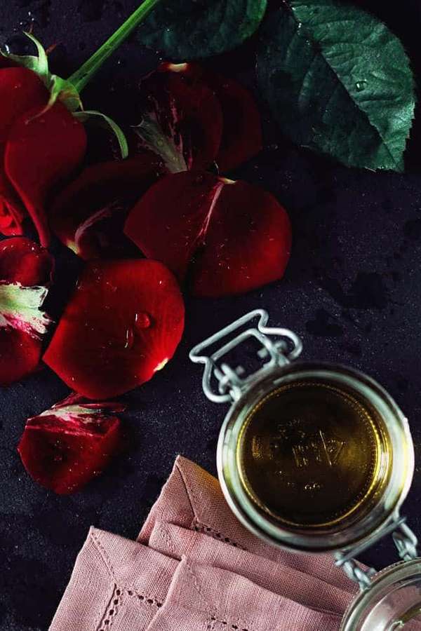 Pétalos de rosa rojos y hojas verdes junto a un frasco de vidrio con líquido dorado sobre superficie oscura con paño rosa. — Aceite de Rosa Fondos de pantalla