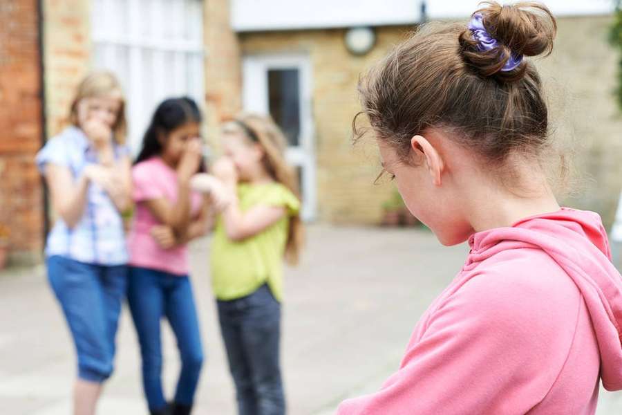 A girl in a pink hoodie stands alone as three other girls laugh behind her. — Actions Picture
