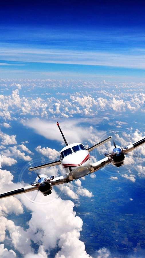 A white propeller plane flying above fluffy clouds in a clear blue sky. — Airplane Android Wallpaper