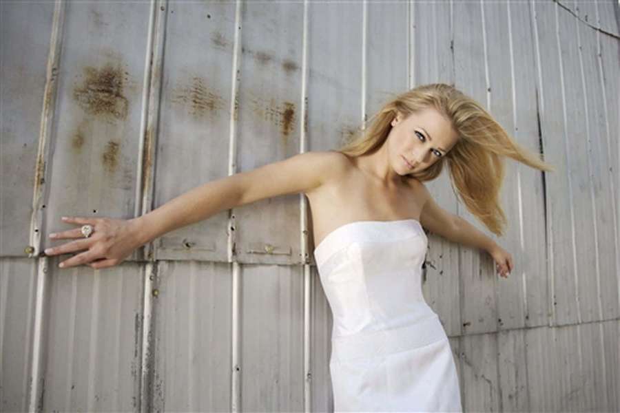 Blonde woman in white dress leaning against rusty metal wall with wind in her hair. — Aj Cook Wallpaper