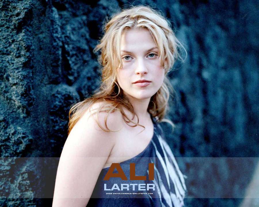 A blonde woman in a dark top stands before a rocky cliff, looking directly at the camera. — Ali larter Background