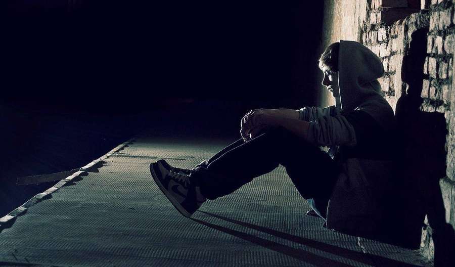 Person in hoodie sitting alone against brick wall in dark alley with single light beam. — Alone Boy Wallpaper
