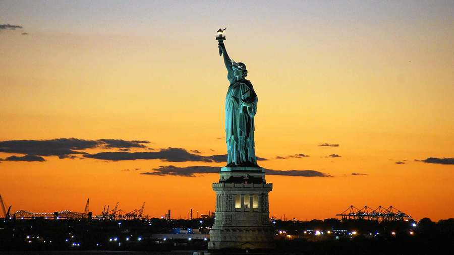 Statue of Liberty illuminated at sunset with harbor cranes and city lights in background. — America Background