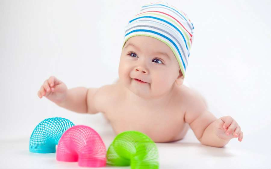 Un bebé feliz con un gorro a rayas juega con juguetes primaverales coloridos sobre una superficie blanca. — Amor de bebé Fondos de pantalla