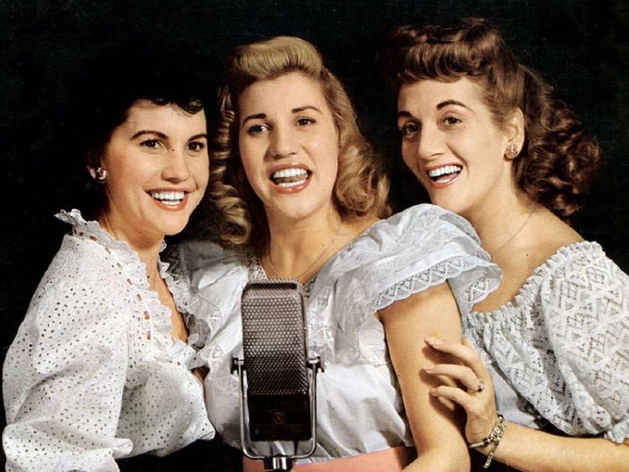 Kolme vintage-naislaulajaa valkoisissa pitsimekoissa hymyilemässä klassisen mikrofonin ympärillä. — Andrews Sisters Taustakuvat