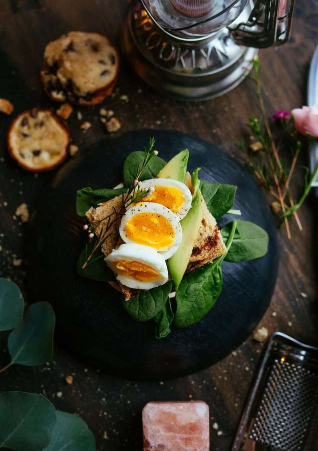 A gourmet breakfast sandwich with egg avocado and spinach on a dark plate with cookie crumbs and herbs. — Android Food Background