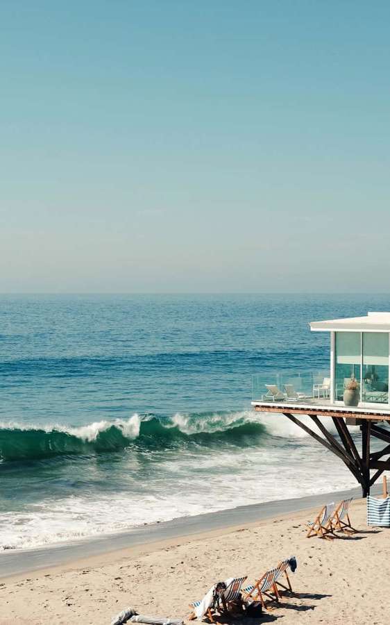 Modern beach house on stilts with ocean waves and sunlit sand. — Android Malibu Background