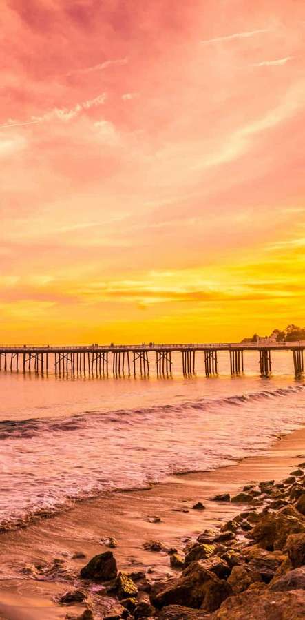 Ein Holzsteg bei Sonnenuntergang mit rosa und orangefarbenem Himmel, der sich im ruhigen Wasser und felsigen Ufer spiegelt. — Android Malibu Hintergrundbild