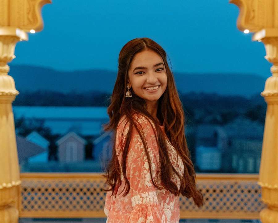 A young woman in a floral dress smiles on a golden balcony at twilight. — Anushka Sen Wallpaper
