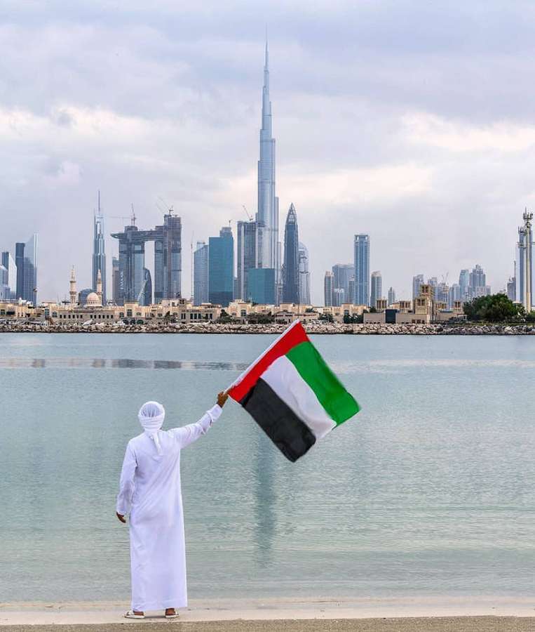Henkilö valkoisessa kandurassa heiluttaa UAE:n lippua Dubain veden äärellä, taustalla Burj Khalifan siluetti. — Arabimies Taustakuvat
