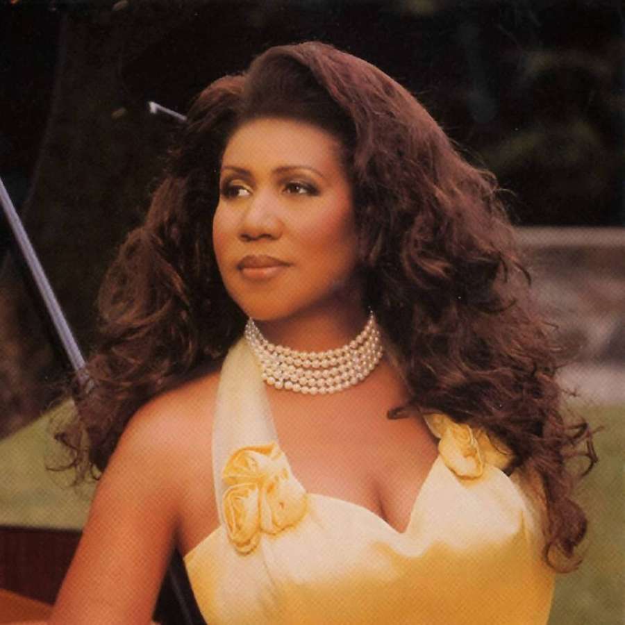 Woman with curly hair wearing yellow dress and pearl necklace outdoors. — Aretha Franklin Wallpaper