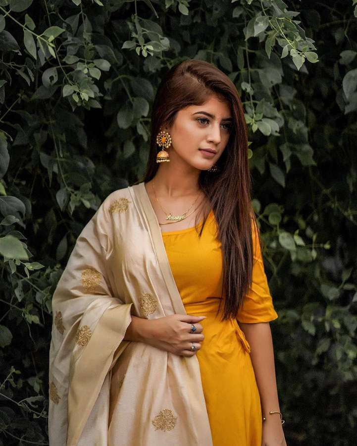 Woman in yellow dress and cream shawl with gold embroidery standing among green foliage. — Arishfa Khan Wallpaper
