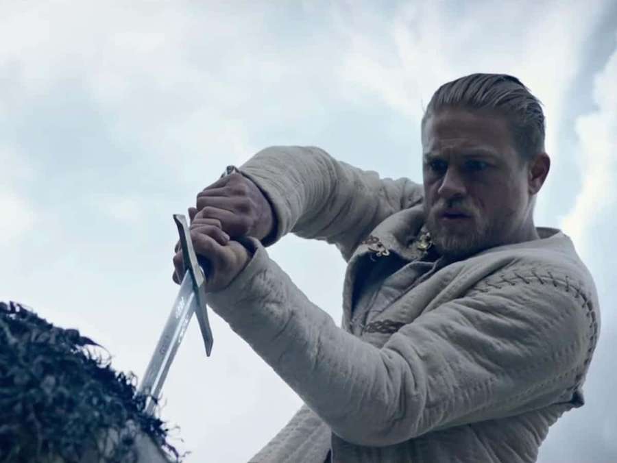 A man in medieval clothing swings a sword with determination against a cloudy sky. — Arthur Background
