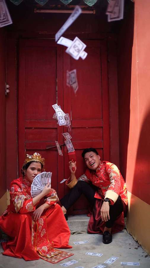 A happy wedding couple in red traditional clothes surrounded by falling paper money. — Asians Background