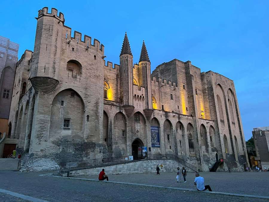 People relax near a historic castle at dusk with warm lights glowing on stone walls. — Avignon Wallpaper