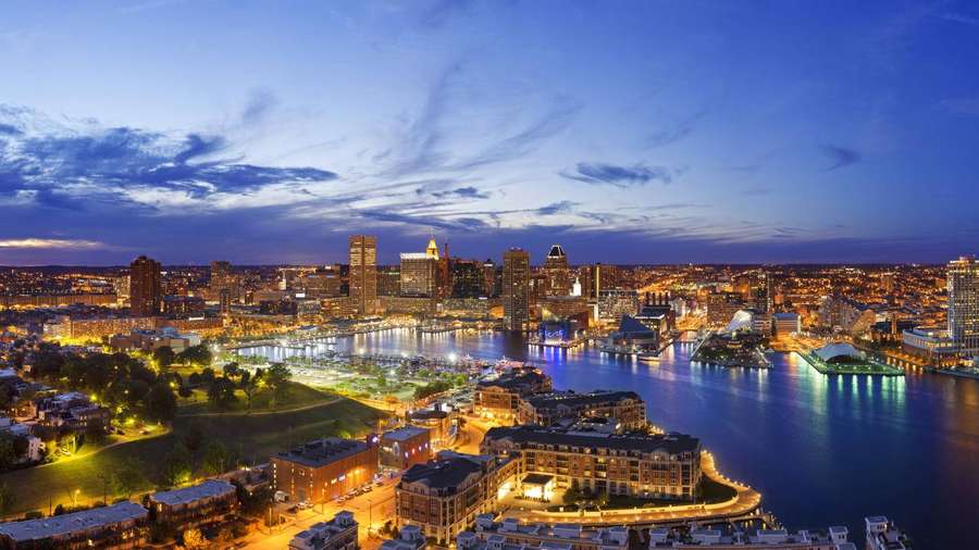 A city skyline at dusk with illuminated buildings and a river reflecting the lights. — Baltimore Wallpaper