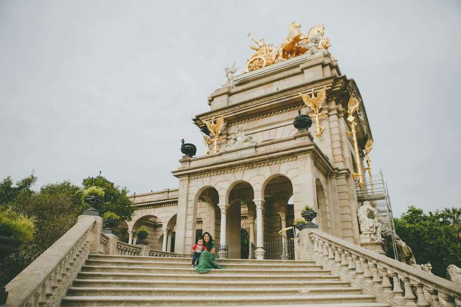 A woman and child sitting on wide stone steps in front of a grand ornate building with golden statues. — Barcelona Background