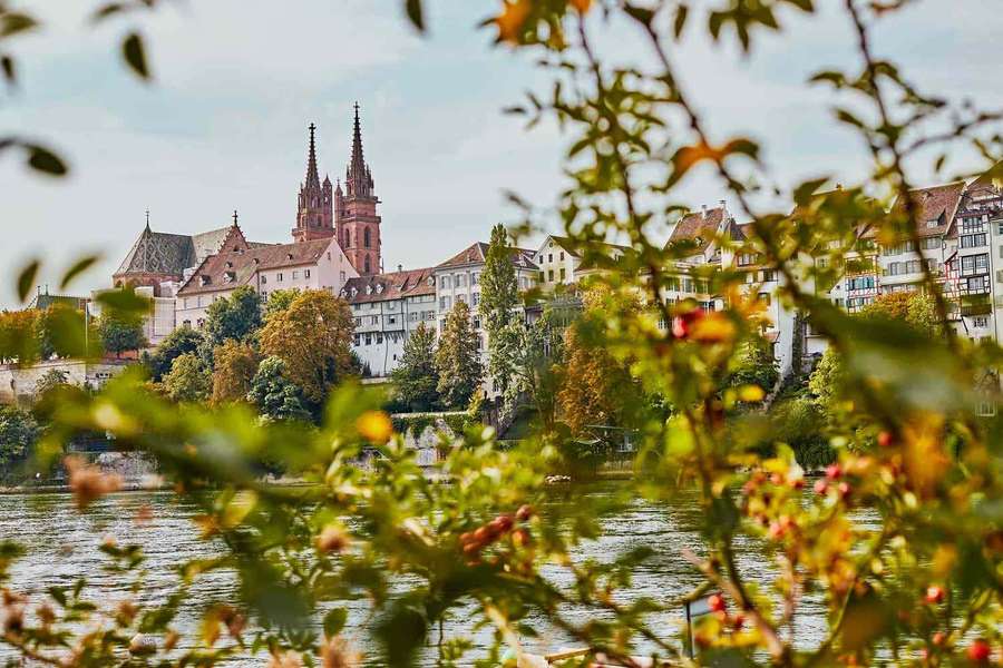 Autumn leaves frame a river view of a historic town with a red brick cathedral. — Basel Wallpaper