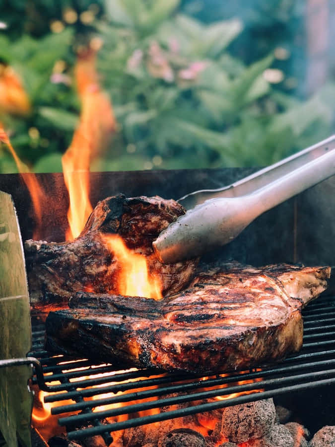 Bistec a la parrilla chisporroteando sobre llamas abiertas en una parrilla de carbón con pinzas ajustándolo. — Bbq Imágenes