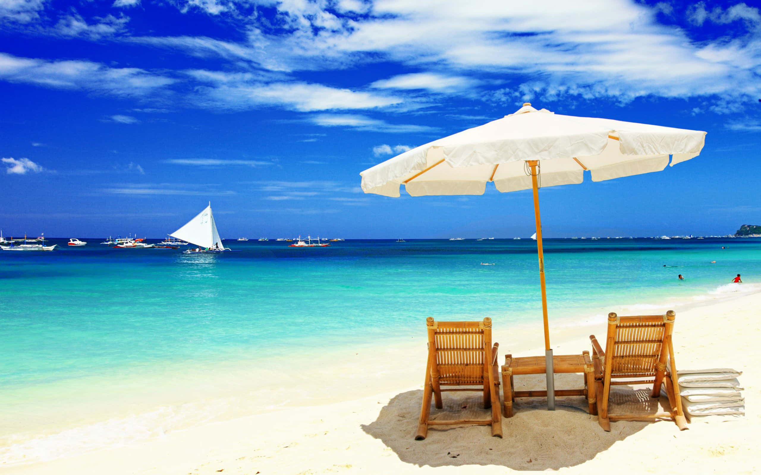 Two wooden chairs under a white umbrella on a tropical beach with sailboats and turquoise water. — Beach Scene Wallpaper