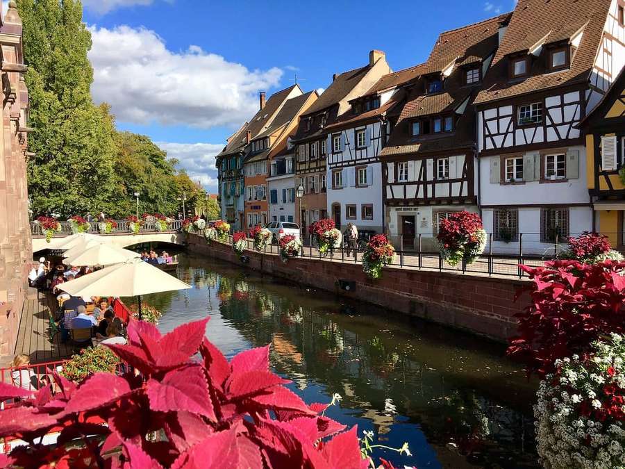 Colorful half-timbered houses line a canal with red flowers and outdoor seating under a blue sky. — Beautiful City Wallpaper