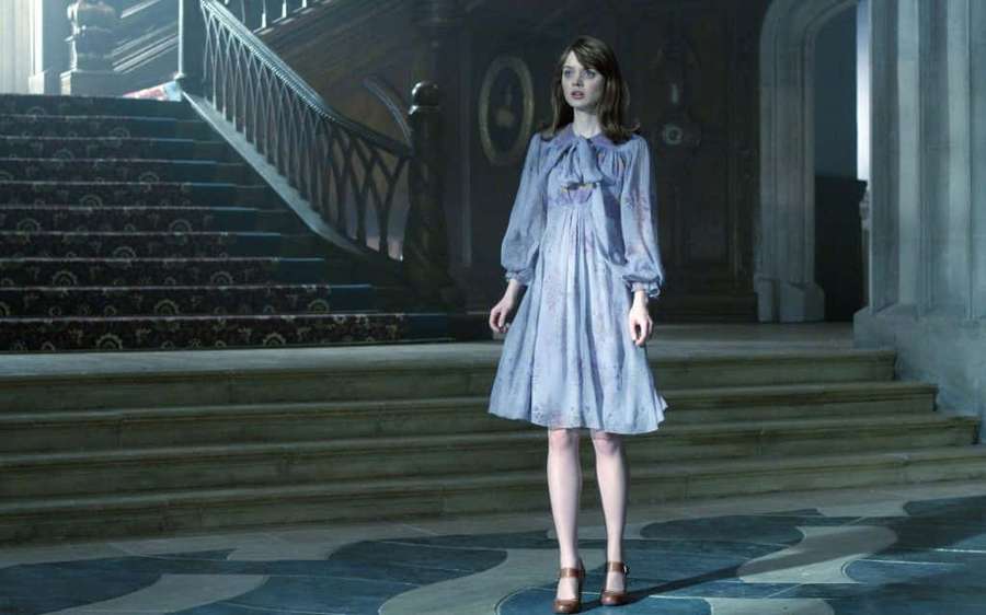 A woman in a blue dress stands in a grand mansion foyer with ornate stairs. — Bella Heathcote Wallpaper
