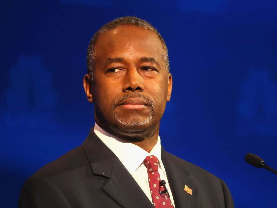 A man in a suit and tie speaking at a podium with a blue background. — Ben Carson Wallpaper
