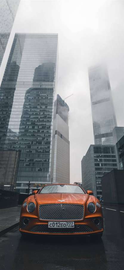 Orange Bentley luxury car parked on wet city street with tall glass buildings in foggy background. — Bentley iPhone Wallpaper