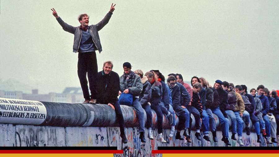 Folla di persone sedute e in piedi sul Muro di Berlino con un uomo che alza le braccia in festa. — Berlin Wall Sfondi