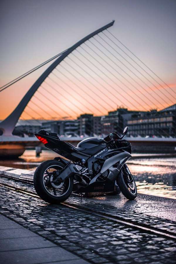 Moto negra estacionada sobre adoquines mojados cerca de puente moderno al atardecer. — Bicicletas Fondos de pantalla
