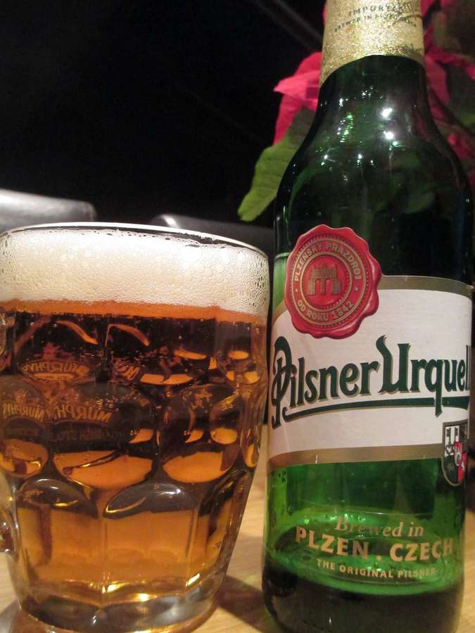 Pilsner Urquell Bierflasche und Glas auf einem Holztisch in einer schwach beleuchteten Umgebung. — Bierflasche Wallpaper