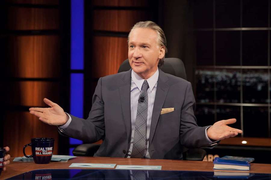 Man in suit gesturing on talk show set with city lights behind him. — Bill Maher Wallpaper