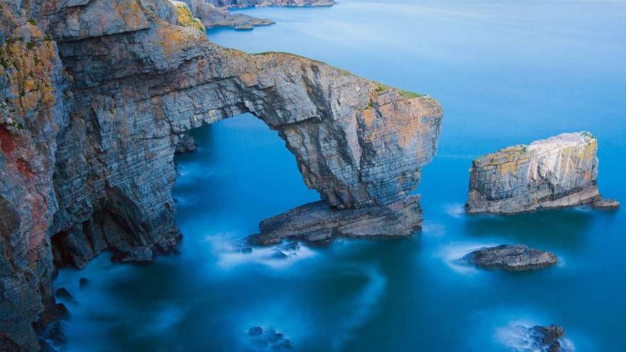 A natural rock arch over calm blue ocean water with misty waves. — Bing Background