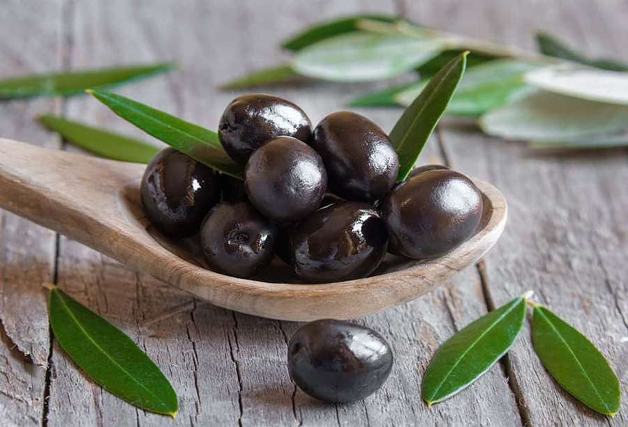 Black olives in wooden spoon with olive leaves on weathered wood. — Black Olives Wallpaper