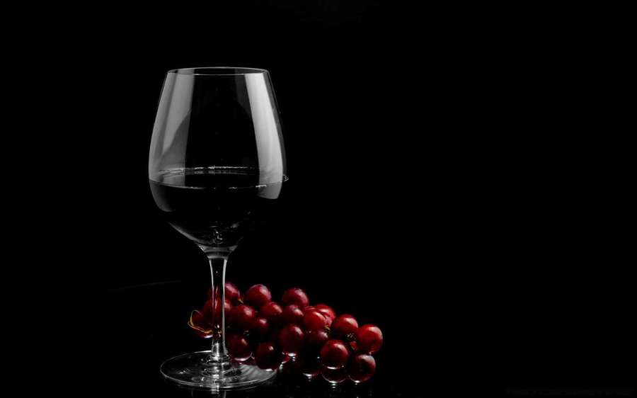 A wine glass and red grapes on a black surface with dramatic lighting. — Black Wine Wallpaper