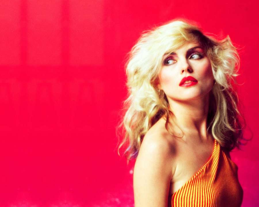 Blonde Frau in orangefarbenem gestreiftem Oberteil vor rotem Hintergrund, die nach oben schaut — Blondie Hintergrundbild