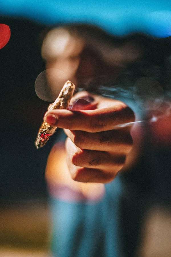 Hand holding lit cigarette with smoke rising in night bokeh lights. — Blunt Wallpaper
