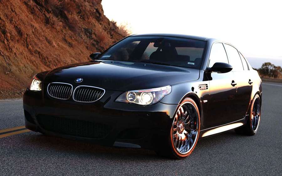 Black BMW M5 sedan with custom wheels on a winding road at sunset. — Bmw M5 Wallpaper