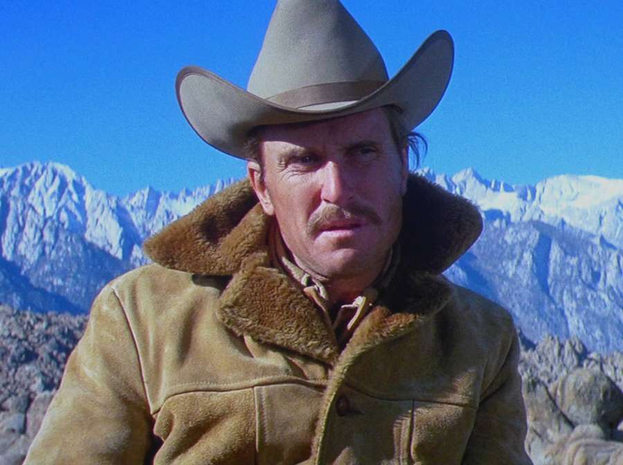 Ein Cowboy in einem pelzgefütterten Mantel und Hut steht vor schneebedeckten Bergen. — Bob Duvall Wallpaper