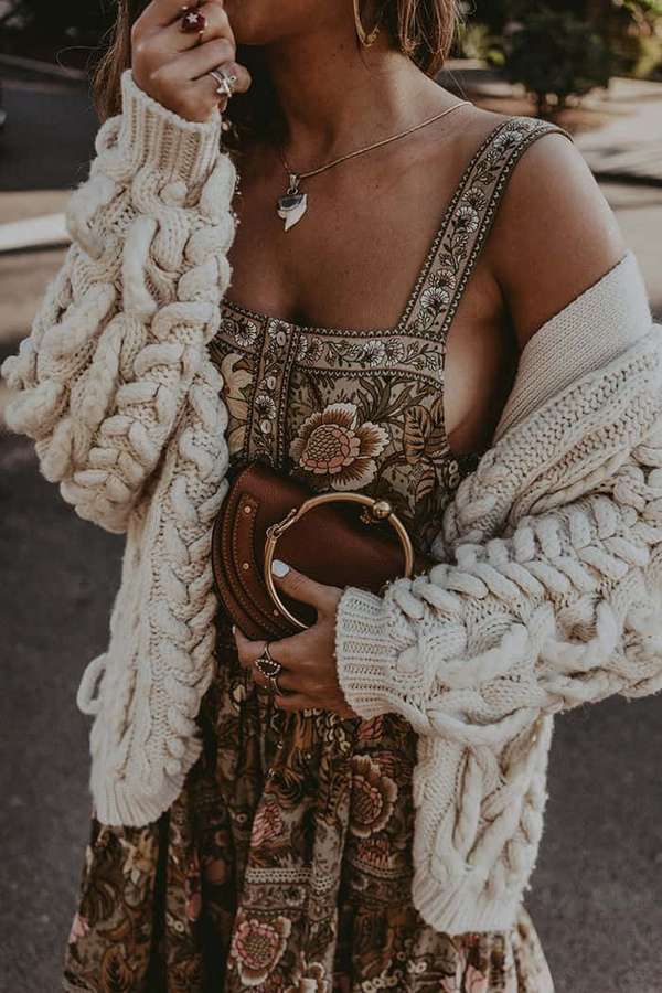 Frau in geblümtem Kleid und cremefarbener Strickcardigan hält braune Clutch im Freien. — Boho Bilder