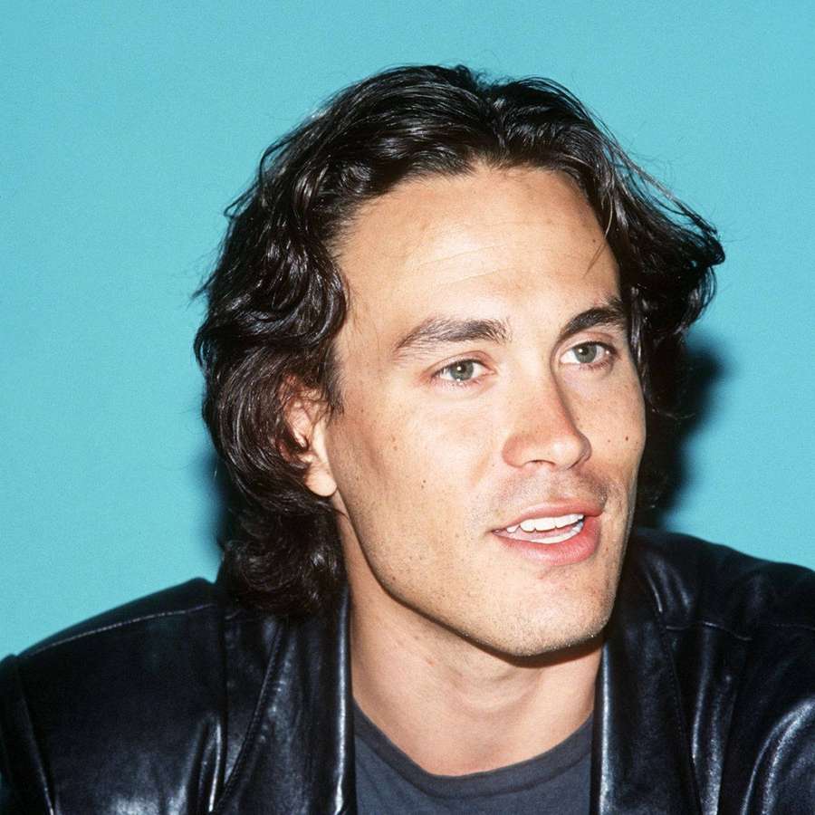 Man with dark hair and green eyes wearing a black leather jacket against teal background. — Brandon Lee Wallpaper