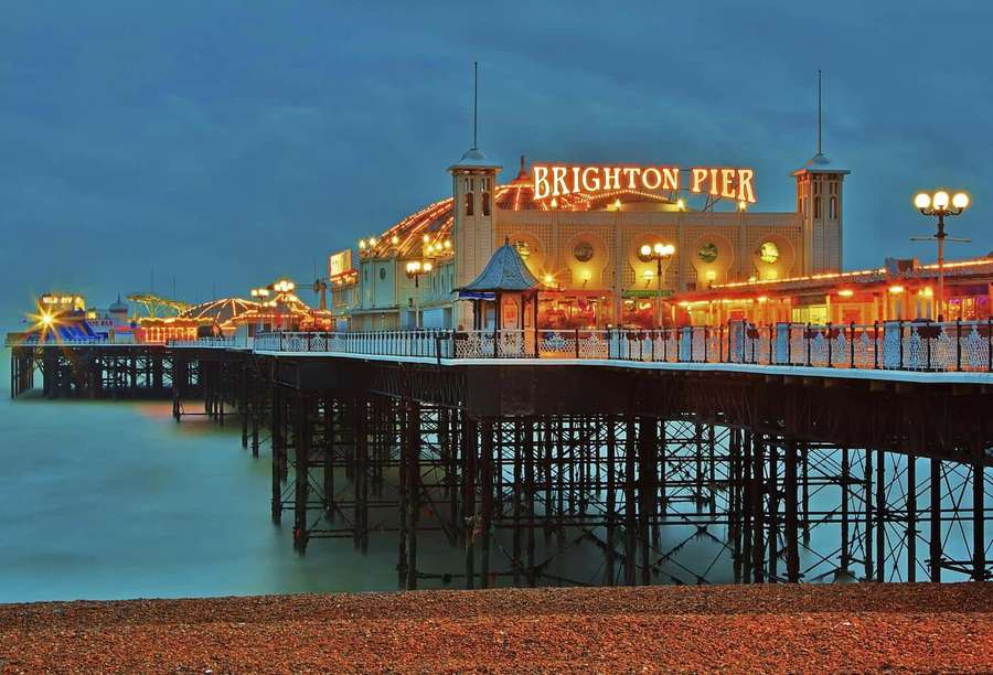 Brighton Pier illuminated at dusk with lights reflecting on water and pebbles. — Brighton Pier Wallpaper