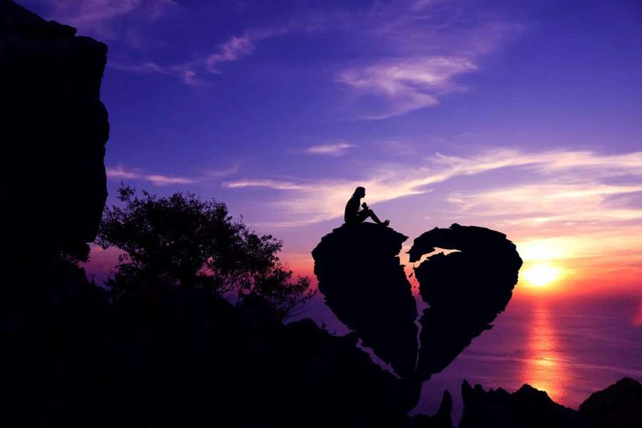 Silhouette of a person sitting on a broken heart rock at sunset over the ocean. — Broken Picture