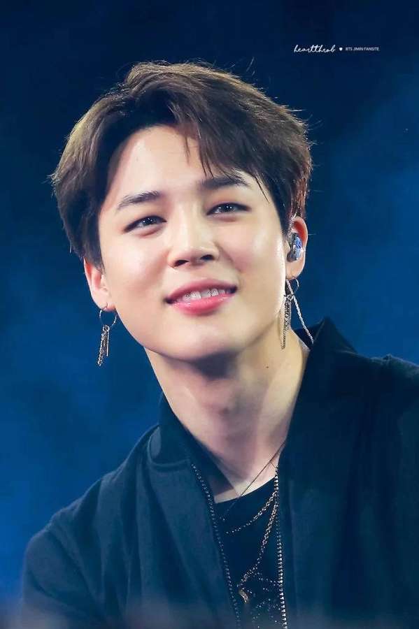 Junger Mann lächelt mit Ohrringen und schwarzer Jacke auf der Bühne unter blauen Lichtern. — Bts Jimin Bilder