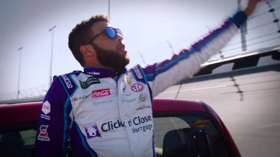 Piloto de corrida acenando para a multidão ao lado de seu carro vermelho em pista ensolarada. — Bubba Wallace Papéis de Parede
