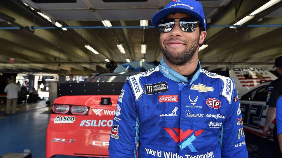 NASCAR-Fahrer in blauem Rennanzug lächelt in Garage neben rotem Rennwagen. — Bubba Wallace Hintergrundbild
