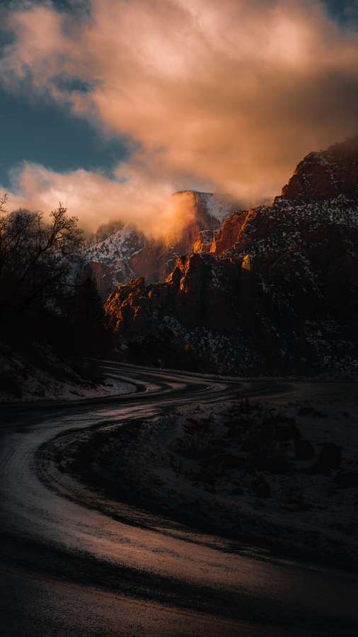 Carretera sinuosa a través de montañas nevadas al atardecer con luz dorada abriéndose paso entre las nubes. — Camino Fondos de pantalla