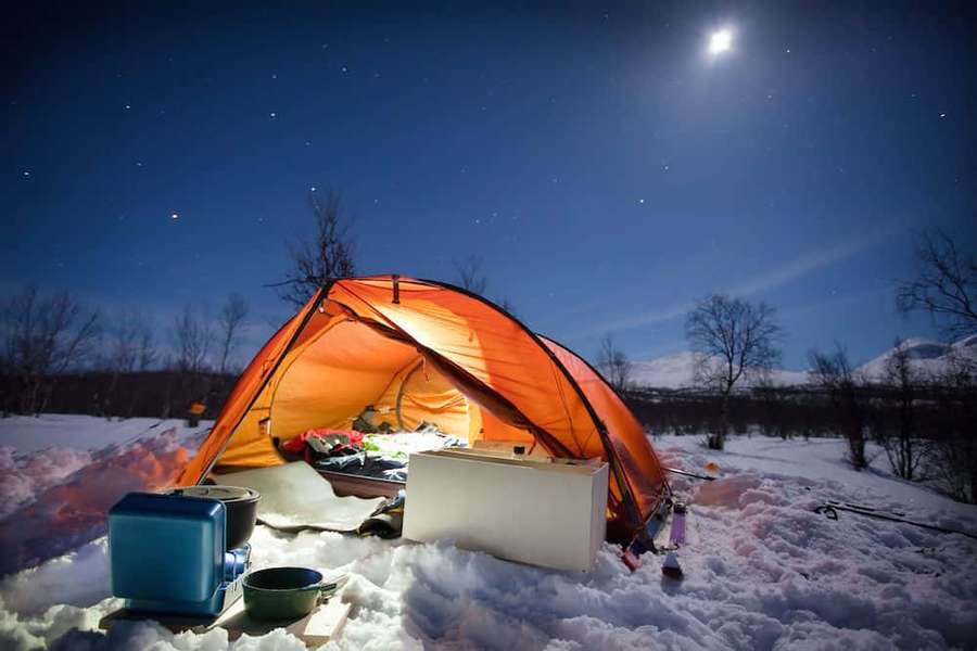 Una tienda naranja cálidamente iluminada en un campo nevado bajo un cielo nocturno estrellado con una luna brillante. — Camping de invierno Fondos de pantalla